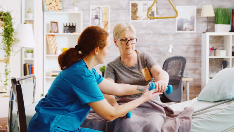 revealing-shot-female-nurse-helping-elderly-woman-recover-her-muscles-after-accident-she-is-lying-hospital-bed-retirement-home